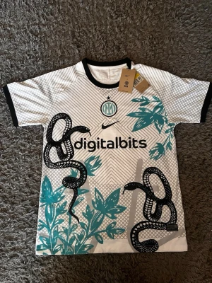 Inter Milan Nike matchtröja - Graphic design - Snygg Inter Milan matchtröja från Nike i vitt med svarta och turkosa detaljer. Tröjan har ett unikt ormmönster och grafiska blad, svart krage och ärmslut samt klubbmärke och sponsortryck framtill. Perfekt för dig som älskar fotboll och vill sticka ut på planen.