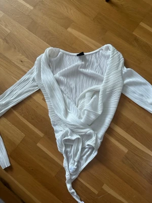 Vitt plisserat omlottbody - Säljer ett vitt body med plisserat tyg och omlottdesign. Bodyn har långa ärmar och djup v-ringning framtill. Materialet är glansigt och stretchigt.