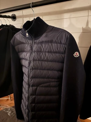 Mörkblå cardiganjacka från Moncler - Snygg mörkblå cardiganjacka från Moncler med quiltad framsida och mjuka stickade ärmar. Dragkedja framtill och hög krage. Perfekt nu till våren när de börjar bli lite varmare. 