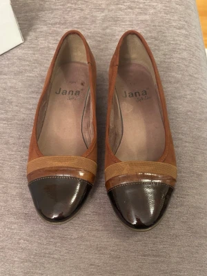 Bruna ballerinaskor från Jana - Snygga ballerinaskor från Jana i brunt mockaliknande material med lackad mörkbrun tå och dekorativ beige rem över tån. Skorna har låg klack och klassisk rundad form. Perfekta för dig som gillar stilrena och tidlösa skor.