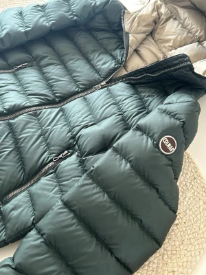 Grön  pufferjacka från Colmar - Säljer en grön pufferjacka från Colmar med snygga horisontella sömmar och två praktiska fickor med dragkedja. Jackan har en rund Colmar-logga på ärmen och är tillverkad i polyester. Perfekt för kalla dagar och har en modern, normal passform.