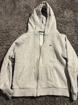 Grå zip-hoodie från Lacoste barn - Snygg grå hoodie med dragkedja från Lacoste. Klassisk krokodillogga på bröstet, två fickor fram och ribbade muddar. Barn storlek och öppen för diskutering av pris!