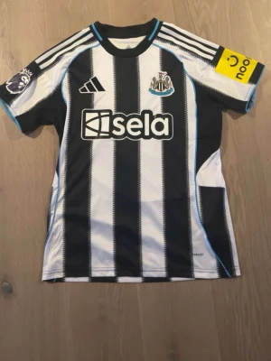 Newcastle Jersey Bruno G. - En helt ny Newcastle tröja beställd direkt från deras hemsida. Den passade mig dock inte och därför jag säljer. Svartvit randig Newcastle United matchtröja från Adidas med trycket 'Bruno G. 39' på ryggen. Tröjan har Premier League-märke på ärmen, sponsorloggor och blå detaljer vid krage och ärmslut. Perfekt för fotbollsfans som vill sticka ut på läktaren.