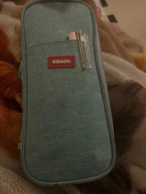 Grön pennfodral från Angoo - Praktiskt pennfodral i grön textil från Angoo med dragkedja och flera fack för pennor och småprylar. Fodralet har en stilren, avlång form och ett litet rött märke på framsidan. Perfekt för att hålla ordning på dina skrivsaker.