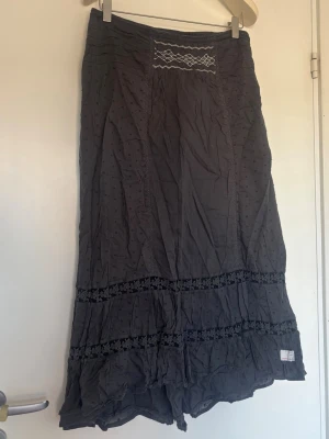 Mörkgrå långkjol med spetsdetaljer - Säljer en mörkgrå långkjol med volanger och fina spetsdetaljer. Kjolen har små prickar i tyget och dekorativa band med blommönster. Storlek 1 vilket motsvarar S