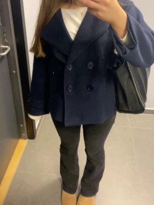 Mörkblå skepparkavaj med bred krage - Säljer en stilren mörkblå skepparkavaj med bred krage och dubbelknäppning framtill. Jackan har en loose passform och är tillverkad i ullmaterial som ger en lyxig och varm känsla. Perfekt för dig som gillar klassisk och cool stil.