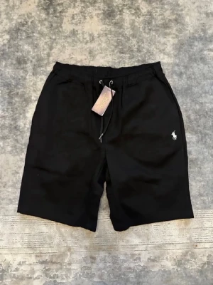 Svarta shorts från Polo Ralph Lauren - Svarta shorts från Polo Ralph Lauren med dragsko i midjan och klassisk logga broderad på vänster ben. Shortsen har sidofickor och en bakficka, perfekt för chill dagar. Tillverkade i mjukt bomullsmaterial för skön känsla.