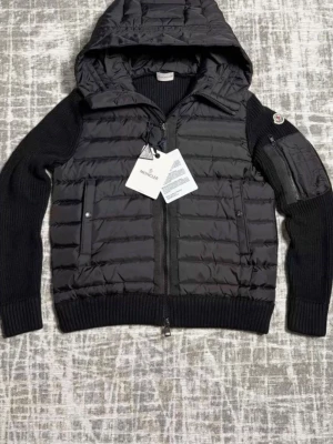 Svart pufferjacka från Moncler - Snygg svart pufferjacka från Moncler med quiltad front och huva. Jackan har stickade ärmar och muddar, dragkedja framtill och fickor med knappstängning. Moncler-logga på ärmen och extra ficka med dragkedja på armen. Perfekt för kyliga dagar.