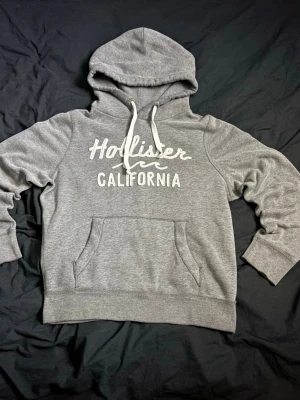 Grå Hollister hoodie med tryck - Fet grå hoodie från Hollister med vit broderad text 'Hollister California' på bröstet.  Mjuk och bekväm bomullsblandning, perfekt för chill dagar eller när du vill ha en avslappnad stil.