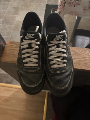Svarta Nike sneakers med vita detaljer - Säljer ett par svarta Nike sneakers med vita kontrastsömmar och vita skosnören. Klassisk låg modell med tydlig Nike-logga på plösen och sidan. Ovandelen är i slätt material och sulan är svart. Perfekta för en clean och sportig stil.