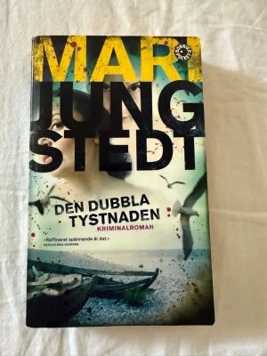 Den dubbla tystnaden av Jungstedt del 7 i Knutas serien - Kriminalroman av Mari Jungstedt i pocketformat. Omslaget har en dramatisk design med stora gula och svarta bokstäver, havsmotiv och fåglar. Boken är tryckt på papper och har en spännande handling som utspelar sig på Gotland. Del 7 i Knutas serien