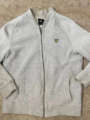 Ljusgrå zip hoodie från Lyle & Scott - Ljusgrå hoodie med dragkedja från Lyle & Scott. Klassisk modell utan huva, med ribbade muddar och två fickor framtill. Broderad gul logga på bröstet. Tillverkad i mjukt bomullsmaterial som känns skönt mot huden.