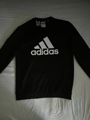 Svart långärmad topp från Adidas - Svart långärmad topp från Adidas med stor vit logga framtill. Klassisk rund halsringning och ribbade muddar vid ärmslut och nederkant. Tillverkad i mjuk bomullsmix som känns skön mot huden. Perfekt för en sportig och avslappnad stil.