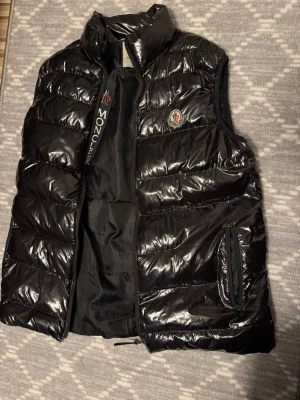 Svart dunväst från Moncler - Snygg svart dunväst från Moncler med glansig finish och klassisk Moncler-logga på bröstet. Västen har hög krage, dragkedja framtill och två sidofickor med dragkedja. Perfekt för lager-på-lager och streetstyle. 