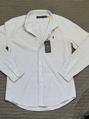 Randig skjorta från Ralph Lauren - Säljer en klassisk vit skjorta med tunna blå ränder från Ralph Lauren. Skjortan har lång ärm, knappar framtill och ikonisk logga broderad på bröstet. Perfekt för dig som gillar stilrena och tidlösa plagg.