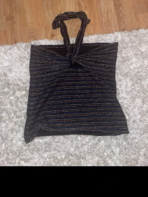 Glittrig svart tubtopp med knyt - Säljer en glittrig svart tubtopp med tunna, färgglada ränder och knyt framtill. Toppen är axelbandslös och har en bandeauformad passning som ger en snygg och trendig look. Perfekt för dig som vill sticka ut lite extra.