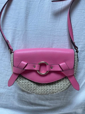 Rosa axelväska med flätad botten - Snygg axelväska i halvmåneform med rosa läderdetaljer och flätad beige botten. Väskan har ett stort silverfärgat spänne framtill och justerbar axelrem. 