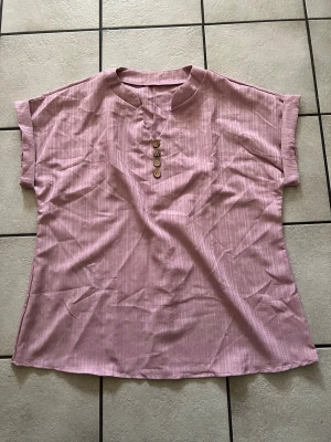 Ljusrosa randig blus med knappar 1XL - Säljer en ljusrosa blus med diskreta ränder och fyra dekorativa knappar framtill. Blusen har korta ärmar och en avslappnad passform med liten ståkrage. Perfekt för dig som gillar en enkel men ändå stilren look.