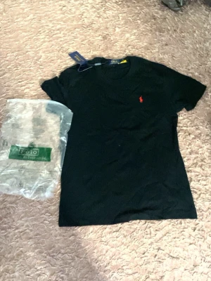 Svart t-shirt från Polo Ralph Lauren - Klassisk svart t-shirt från Polo Ralph Lauren med den ikoniska röda logon broderad på bröstet. T-shirten har rund hals och korta ärmar, perfekt till jeans eller shorts. Enkel och stilren design som funkar till allt.