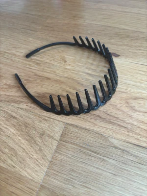 Svart hårband med kammar - Säljer ett svart hårband i plast med inbyggda kammar som håller håret på plats. Perfekt för att skapa en clean look eller hålla undan håret från ansiktet. Enkel och stilren design som passar till många olika frisyrer.