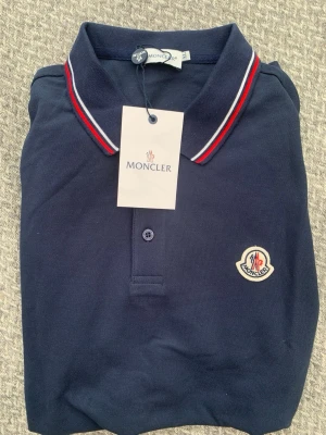 Marinblå pikétröja från Moncler - Snygg marinblå pikétröja från Moncler med klassisk krage och röd-vita ränder. Tröjan har broderad Moncler-logga på bröstet och knäppning framtill. Perfekt för en stilren och sportig look.