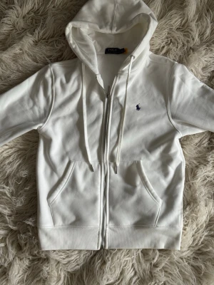 Vit hoodie från Polo Ralph Lauren - Snygg vit hoodie från Polo Ralph Lauren med dragkedja, två fickor framtill och klassisk blå logga på bröstet. Tillverkad i mjukt material och har justerbar huva med snören. Perfekt för en clean och stilren outfit. Passar storlek S.