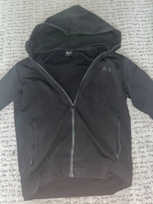 Hugo Boss - Säljer en svart hoodie från BOSS med dragkedja framtill och huva. Diskret BOSS-logga på bröstet och längs ärmarna, samt vita detaljer på ärmarna. Två fickor framtill och ribbade muddar. Perfekt för en chill och stilren look.