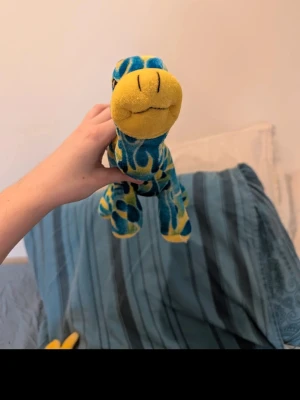 Blågul dinosaurie mjukisdjur - Säljer ett mjukisdjur i form av en dinosaurie med blå och gula mönster. Huvudet och tassarna är gula medan kroppen har ett blått och gult flammigt mönster. Perfekt som dekoration eller gosedjur. Tillverkad i mjukt tyg.