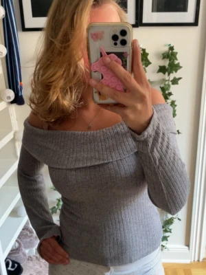 Grå off shoulder ribbad tröja - Trendig grå ribbad tröja med off shoulder-design och långa ärmar. Tröjan har en bred krage som vilar snyggt över axlarna och är tillverkad i ett mjukt, stretchigt material som sitter tajt mot kroppen. Perfekt för dig som vill ha en stilren men ändå cool look.