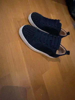 Svarta sneakers med nitar och röd sula - Snygga svarta sneakers med coola nitar på tån och ovandelen. Skorna har en vit sula med röd undersida och svarta skosnören. Insidan är beige och materialet ser ut att vara mocka eller textil. Perfekta för dig som vill sticka ut med din stil.