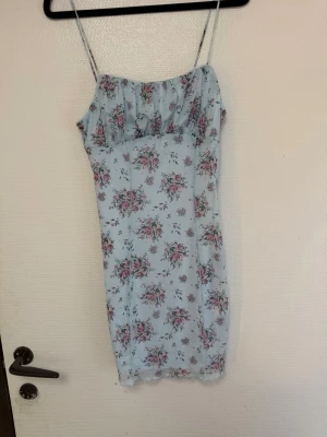 Ljusblå blommig kort klänning - Söt ljusblå kort klänning med tunna axelband och blommigt mönster i rosa och grönt. Klänningen har en figurnära passform och är perfekt för varma dagar. Materialet känns lätt och luftigt, vilket gör den skön att bära under sommaren. ( tänk på jag har hund i hemmet) 