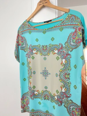 Turkos mönstrad blus med paisley - Snygg turkos blus med färgglatt paisleymönster i rosa, grönt och beige. Blusen har rund halsringning och korta ärmar. Tillverkad i ett lätt och luftigt material som känns skönt mot huden. Perfekt för dig som gillar bohemiska och färgstarka plagg.