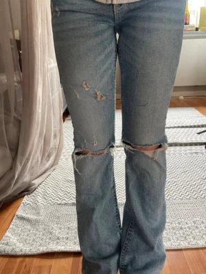 Ljusblå  bootcut jeans - Säljer ett par ljusblå bootcut jeans med slitningar och hål vid knäna. Jeansen har en klassisk femficksmodell och är tillverkade i denim med en avslappnad passform. Perfekta för en trendig och avslappnad stil.