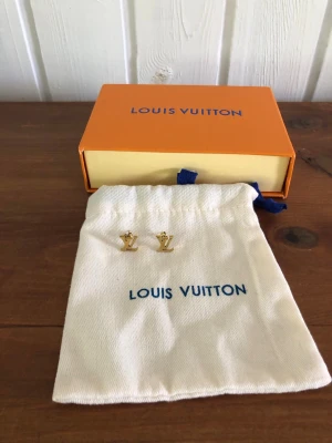 Louis Vuitton guldörhängen LV-logo - Säljer ett par stilrena örhängen från Louis Vuitton i guldfärgad metall. Örhängena har den ikoniska LV-logotypen och kommer med originalpåse och ask. Perfekta för dig som vill ha en exklusiv accessoar med lyxig känsla.
