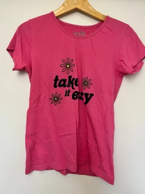 Rosa t-shirt med tryck från Sfera - Snygg rosa t-shirt från Sfera med svart text 'take it easy' och blommor på framsidan. T-shirten har rund halsringning och korta ärmar. Perfekt för en avslappnad och trendig look. Passar en liten M eller vanlig S