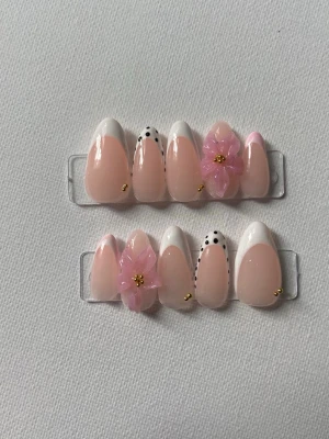 Press-on naglar med blommor och prickar - Snygga press-on naglar i mandelform med ljusrosa bas, vita tippar, svarta prickar och 3D-blommor i rosa med gulddetaljer. Perfekt för dig som vill ha en unik och trendig nageldesign