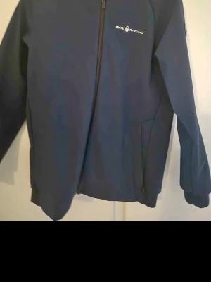 Marinblå vindjacka från Sail Racing - Snygg marinblå vindjacka från Sail Racing med dragkedja framtill och diskret logga på bröstet. Jackan har en minimalistisk design, två sidofickor och är tillverkad i ett lätt, vindtätt material som passar perfekt för blåsiga dagar.