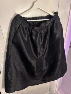 Svart kjol med glansig finish från Stockh LM  - Säljer en svart kjol från Stockh LM med glansig finish. Kjolen har en enkel och klassisk design med resår i midjan och lätt utställd passform. Perfekt att styla med både sneakers och boots.