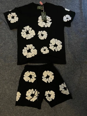 Svart Denim Tears Set - Svart t-shirt från Palm Angels med stora vita blommönster över hela plagget. T-shirten har rund hals och korta ärmar, perfekt för en avslappnad och trendig look. Materialet känns mjukt och bekvämt mot huden.Finns även i andra färger. Storlek S,M och L