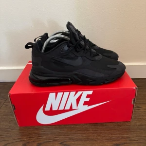 Nike Air270 React svarta sneakers - Nike Air270 React sneakers i helsvart med chunky sula och synlig Air-enhet i hälen. Skorna har en sportig design med mesh och syntetmaterial, samt svarta detaljer och logga på sidan. Perfekta för dig som gillar streetwear och en clean look.