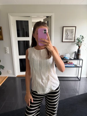 Vit ärmlös blus med knyt i ryggen - Säljer en söt vit ärmlös blus med dekorativa vertikala spetsdetaljer framtill och knyt i ryggen. Blusen har rund halsringning och är lätt figursydd för en snygg passform. Perfekt till både jeans och kjol!