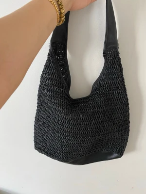 Svart axelväska i flätad design, strand 🏖️ väska - Snygg svart axelväska från NA-KD med flätad design och detaljer i konstläder. Väskan har en mjuk, slouchy form och bred axelrem för skön känsla. Perfekt för dig som gillar trendiga accessoarer med lite edge.