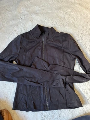 Svart träningsjacka från Gina Tricot - Snygg svart träningsjacka från Gina Tricot med hel dragkedja framtill och hög krage. Tillverkad i ett stretchigt och mjukt material som passar perfekt till träning eller powerwalks. Jackan har långa ärmar och en smickrande passform.