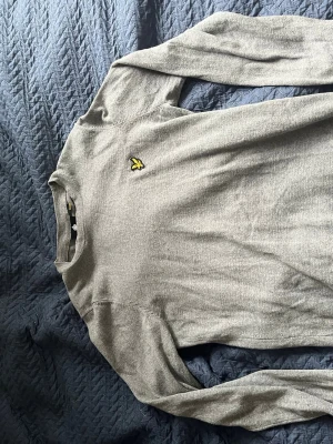 Grå tröja från Lyle & Scott - Snygg grå långärmad tröja från Lyle & Scott med klassisk gul logga på bröstet. Tröjan har rund halsringning och är tillverkad i mjukt material som passar perfekt till jeans eller chinos.