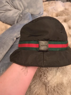 Svart bucket hat - Snygg svart bucket hat med klassiskt grönt och rött band runtom. Hatten är tillverkad i Italien och har en mjuk, lätt glansig yta. Perfekt accessoar för dig som vill sticka ut med en ikonisk och trendig look. Står storlek m men uppskattar en xs-s