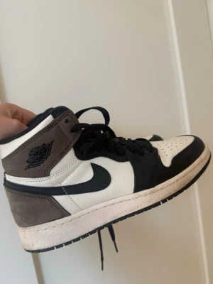 Air Jordan 1 High mocha brun - Säljer ett par Air Jordan 1 High sneakers i svart, vitt och brunt. Skorna har klassisk hög siluett, svart Swoosh och detaljer i mocka och läder. Ikonisk Jordan-logga på sidan och svart snörning. Köpt av Merchsweden. Små i storlek passar 36,5 -37. Använt cirka 4-5 gågner 