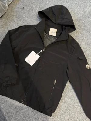 Svart vindjacka från Moncler - Snygg svart vindjacka från Moncler med huva och dragkedja framtill. Jackan har två framfickor med dragkedja och Moncler-logga på ärmen. Tillverkad i lätt och slitstarkt material som passar perfekt för blåsiga dagar.