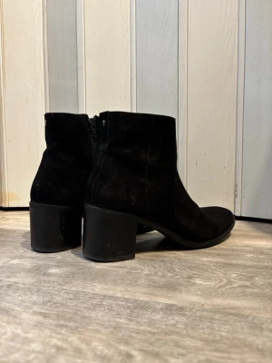 Svarta mockastövletter med klack - Säljer ett par svarta stövletter i mocka med bred klack och dragkedja på insidan. Klassisk och stilren modell som passar till det mesta. Skorna har rundad tå och en stabil klack som ger extra höjd utan att tumma på komforten.