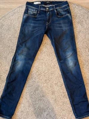 Blå slim fit jeans från Replay - Säljer ett par blå slim fit jeans från Replay med snygga slitningar och kontrastsömmar. Jeansen har klassiska fem fickor, läderpatch bak och coolt mönster på bakfickorna. Perfekt för dig som gillar en modern och avslappnad look.