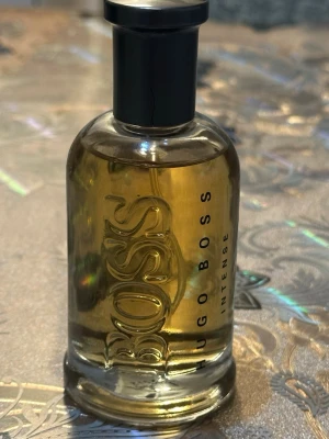 Hugo Boss Bottled Intense parfym - En stilren parfym från Hugo Boss, Bottled Intense, i en genomskinlig glasflaska med svart spraykork. Flaskan har en rundad form och guldgul vätska inuti. Boss-loggan är präglad på framsidan och texten 'Hugo Boss Intense' syns tydligt. Oanvänt 
